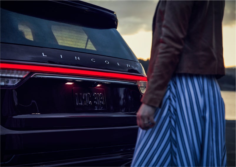 A person is shown near the rear of a 2023 Lincoln Aviator® SUV as the Lincoln Embrace illuminates the rear lights | Griffin Lincoln in Tifton GA